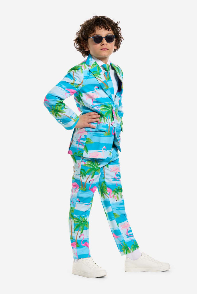 In this image, a kid is wearing a light blue boys’ suit with a tropical print and a white shirt. The suit has a blue print resembling water and pink flamingos and green palm trees. Kid wearing sunglasses and holding a hand on his hip.