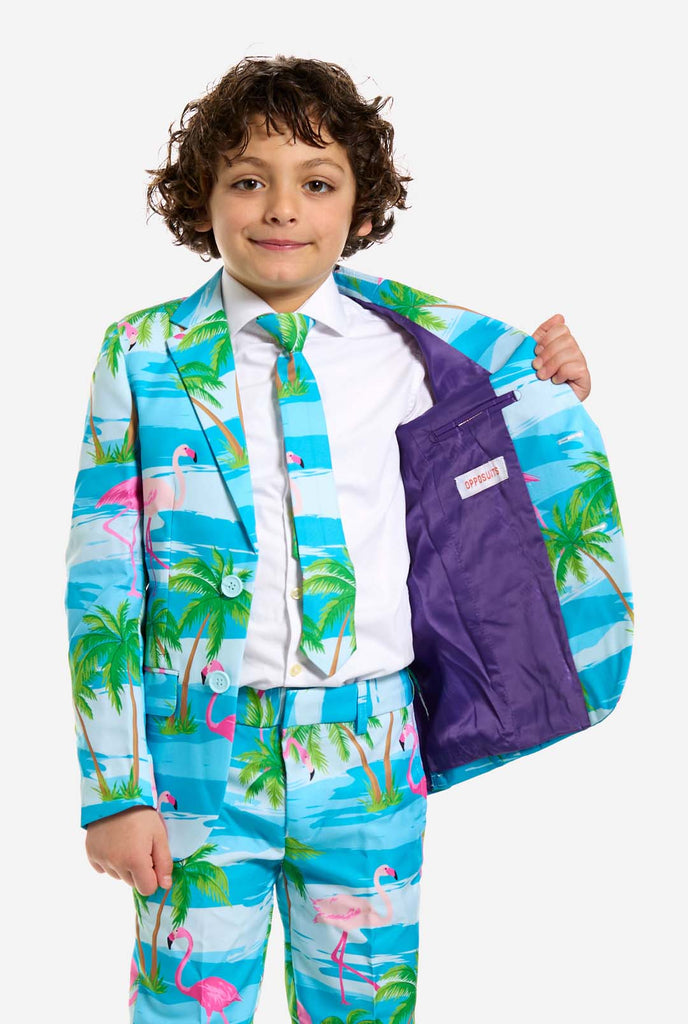 In this image, a kid is wearing a light blue boys’ suit with a tropical print and a white shirt. The suit has a blue print resembling water and pink flamingos and green palm trees. Holding one side of the jacket open.