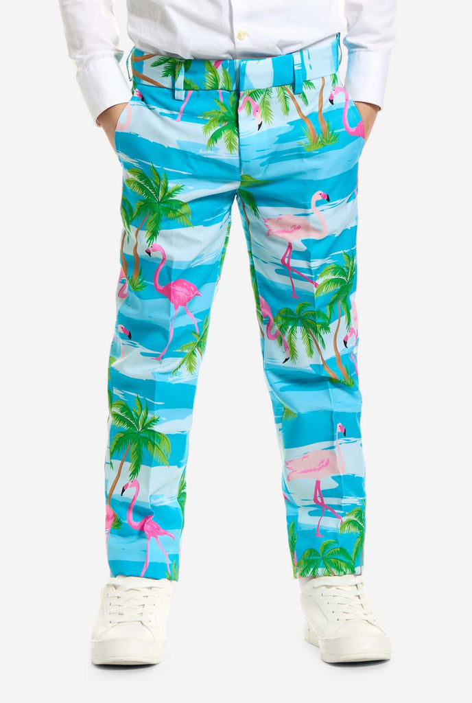 In this image, a kid is wearing a light blue boys’ suit with a tropical print and a white shirt. The suit has a blue print resembling water and pink flamingos and green palm trees. Close up of pants.