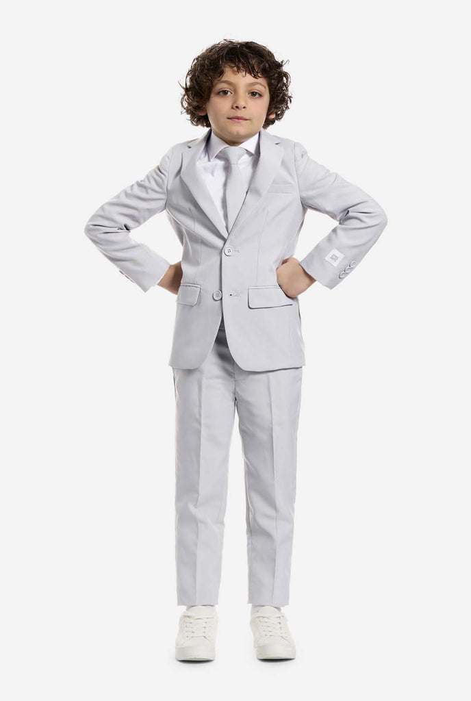 In this image, a kid is wearing a grey formal boys’ suit with a matching tie and a white shirt. The suit consists of a jacket, pants and clip on tie. Holding hands on his hips.