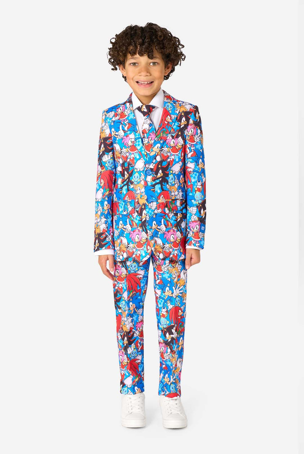 Young boy wearing a vibrant Sonic teen suit featuring colorful prints of Sonic the Hedgehog characters including Sonic, Knuckles, Tails, and Amy, paired with a matching tie and white shirt. Looking in the camera.