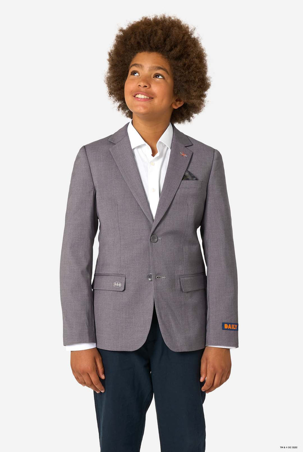 A child smiling while wearing a light grey blazer, white shirt, and dark trousers, looking upward.