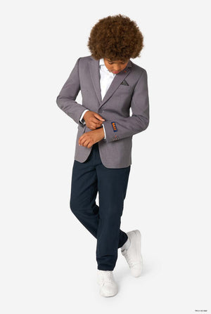 Full-body view of a child in a grey blazer, white shirt, dark trousers, and white sneakers, adjusting the sleeve.
