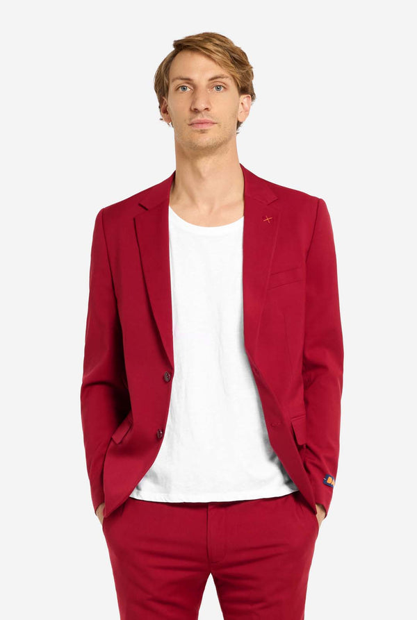 Image with light grey background and man wearing Bright Burgundy men’s suit and white shirt. Suits consists of blazer and pants. Man looking in the camera.