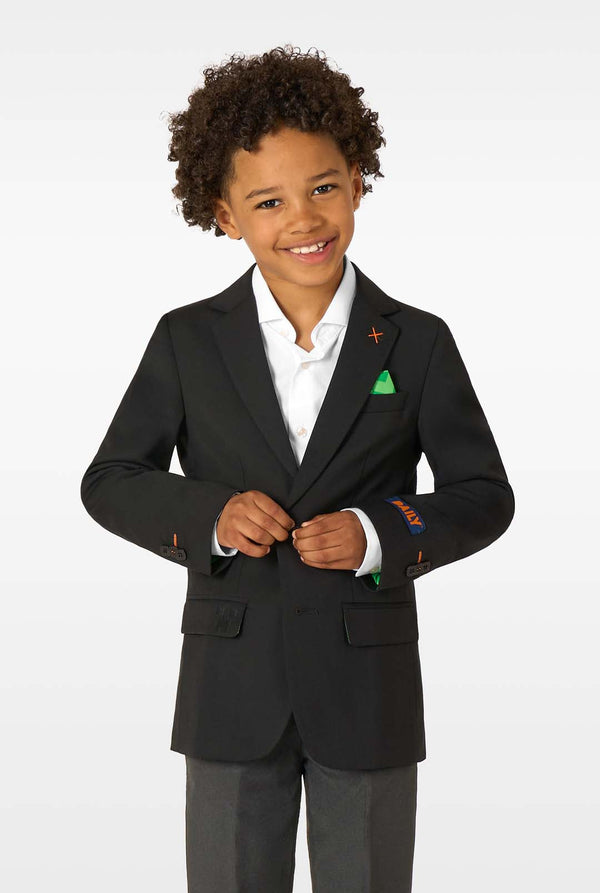 A young boy smiling while wearing a black blazer, white shirt, and grey trousers with a green pocket square.