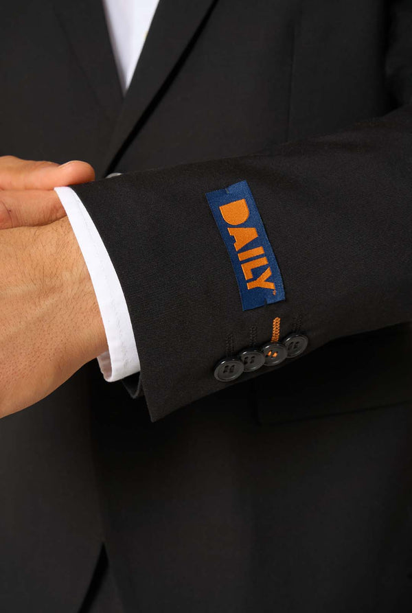 Close-up of the sleeve of a black blazer with a navy and orange “DAILY” label and black button details.