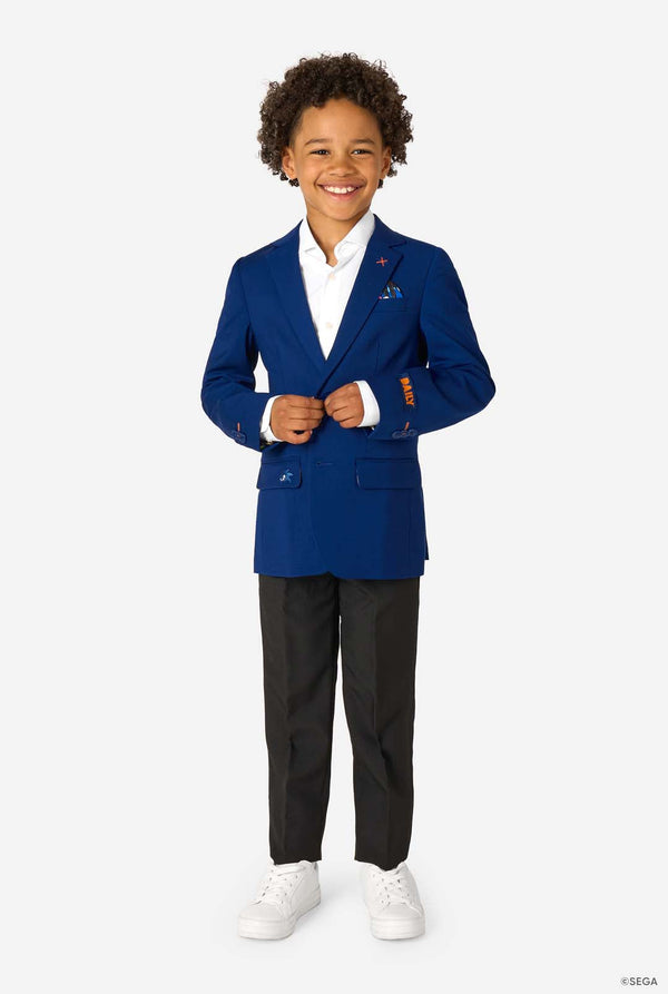 Full-body image of a boy in a blue blazer, white shirt, black trousers, and white sneakers, buttoning his jacket.
