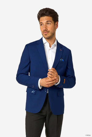 A man wearing a bright blue blazer, white shirt, and black trousers, standing with hands clasped and looking to the side.