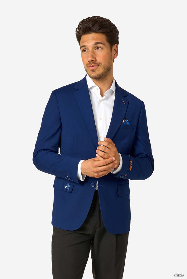 A man wearing a bright blue blazer, white shirt, and black trousers, standing with hands clasped and looking to the side.