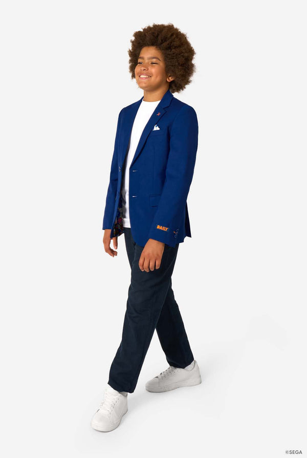 A child walking confidently in a blue blazer, white T-shirt, dark trousers, and white sneakers, smiling.