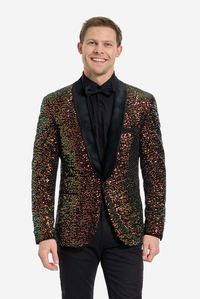 A man is standing and smiling, dressed in a formal, eye-catching outfit. He is wearing a black dress shirt with a black bow tie and a blazer adorned with shimmering amber-colored sequins that reflect light in various shades of gold, copper, and green. The blazer has a satin black shawl collar and front pockets. He pairs this bold jacket with black dress pants, creating a contrast between the sparkly top and the sleek bottom. The background is plain, emphasizing the dazzling effect of the sequined blazer.