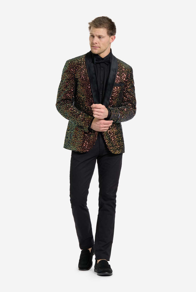 A man is standing and looking side ways dressed in a formal, eye-catching outfit. He is wearing a black dress shirt with a black bow tie and a blazer adorned with shimmering amber-colored sequins that reflect light in various shades of gold, copper, and green. The blazer has a satin black shawl collar and front pockets. He pairs this bold jacket with black pants, creating a contrast between the sparkly top and the sleek bottom. The background is plain, emphasizing the dazzling effect of the sequined blazer.