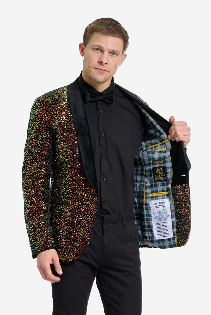 A man is standing dressed in a formal, eye-catching outfit. He is wearing a black dress shirt with a black bow tie and a blazer adorned with shimmering amber-colored sequins that reflect light in various shades of gold, copper, and green. The blazer has a satin black shawl collar and front pockets. He pairs this bold jacket with black dress pants. The background is plain, emphasizing the dazzling effect of the sequined blazer. Holding one side of jacket open.