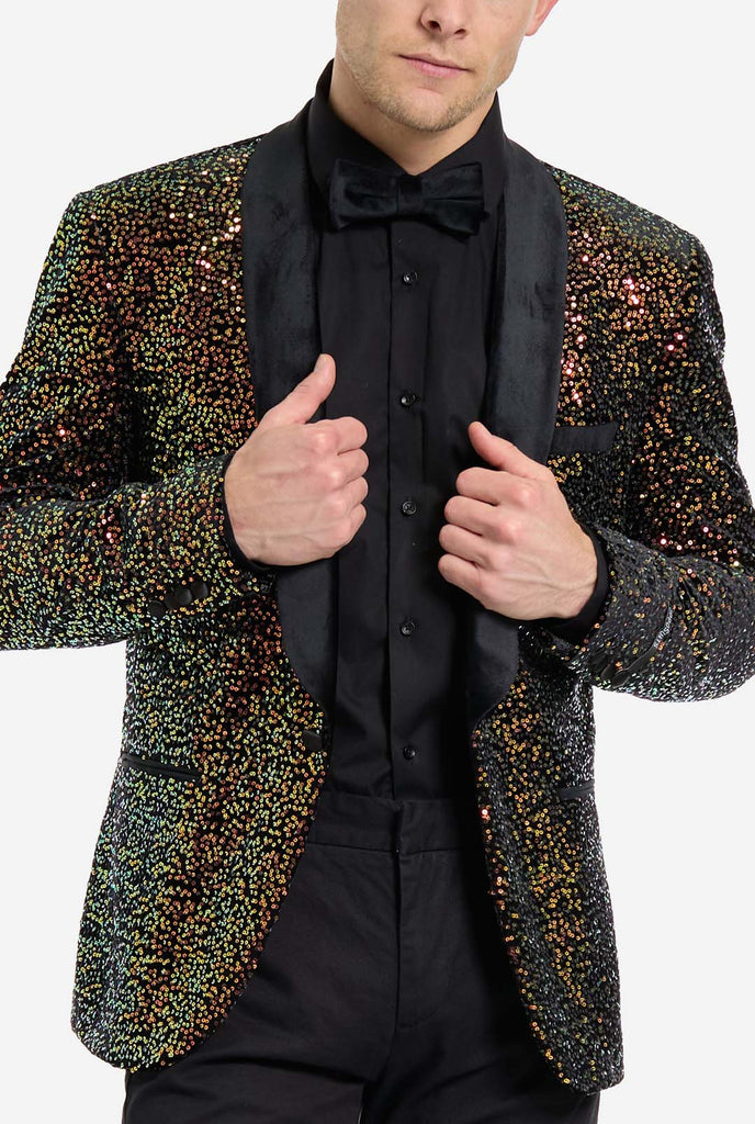 A man is dressed in a formal, eye-catching outfit. He is wearing a black dress shirt and a blazer adorned with shimmering amber-colored sequins that reflect light in various shades of gold, copper, and green. The blazer has a satin black shawl collar. He pairs this bold jacket with black dress pants, creating a contrast between the sparkly top and the sleek bottom. The background is plain, emphasizing the dazzling effect of the sequined blazer. Chest close up.