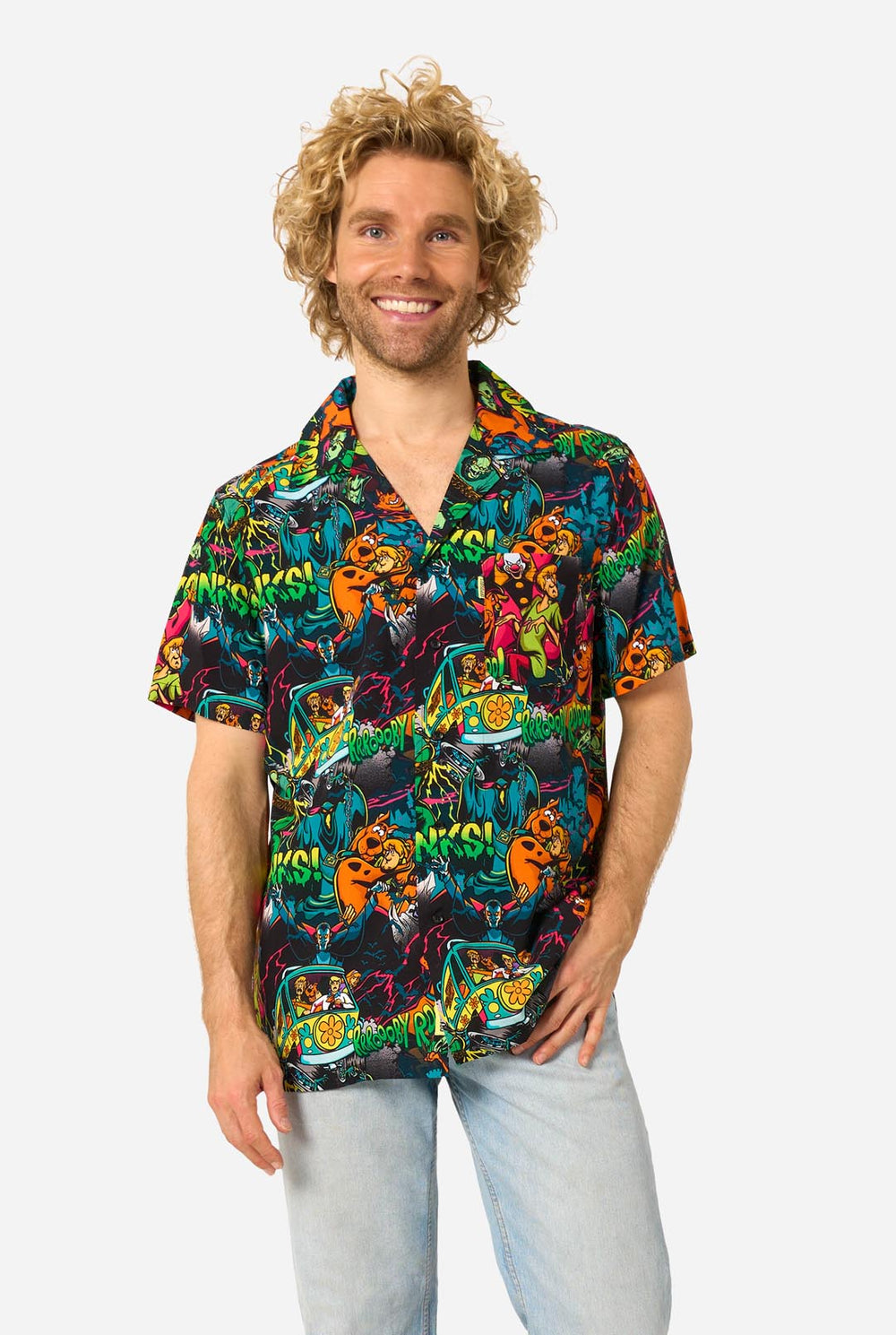 A man with curly blonde hair wearing a short-sleeve Hawaiian shirt with a vibrant, colorful, graphic Scooby-Doo print on a black background, paired with light blue jeans, against a plain light gray background.
