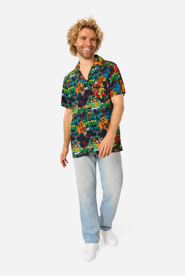 A man with curly blonde hair wearing a short-sleeve Hawaiian shirt with a vibrant, colorful, graphic Scooby-Doo print on a black background, paired with light blue jeans, against a plain light gray background. Full body image.