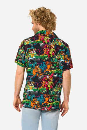 A man with curly blonde hair wearing a short-sleeve Hawaiian shirt with a vibrant, colorful, graphic Scooby-Doo print on a black background, paired with light blue jeans, against a plain light gray background. View from the back.