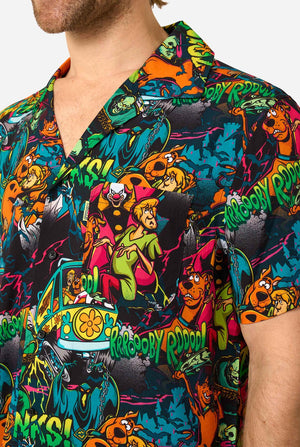 A man with curly blonde hair wearing a short-sleeve Hawaiian shirt with a vibrant, colorful, graphic Scooby-Doo print on a black background, against a plain light gray background. Chest close up.