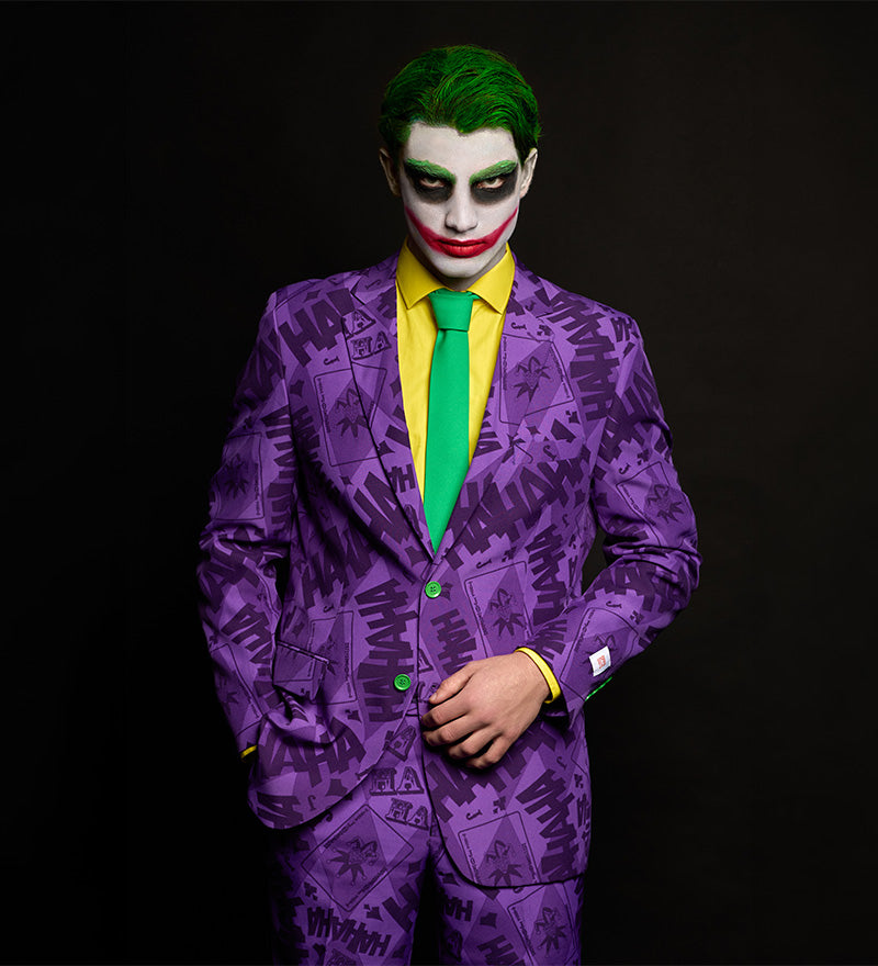 A person wearing a purple suit with a repeating Joker-style print, a bright yellow shirt, and a green tie, styled like the Joker with green hair and white face makeup with red smile. The background is solid black.