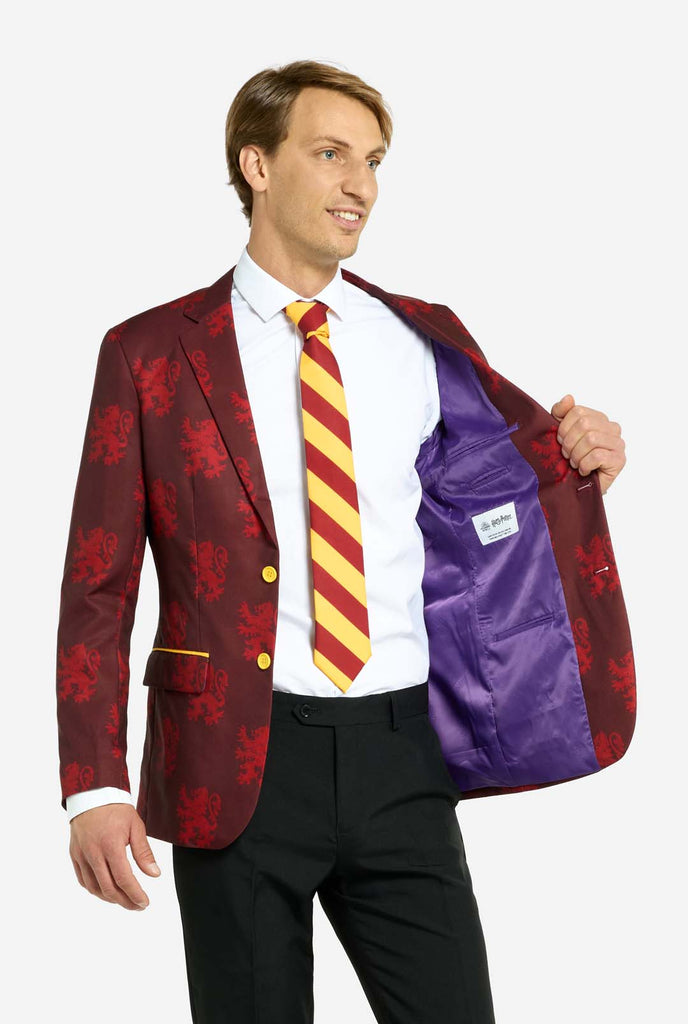 Image with light grey background and man wearing Harry Potter suit, Gryffindor styled. Suit consists of red blazer with Gryffindor logo and light red lions all over print, black pants, and red and yellow diagonal striped tie. Man holding one hand of the blazer open, showing purple inside.   