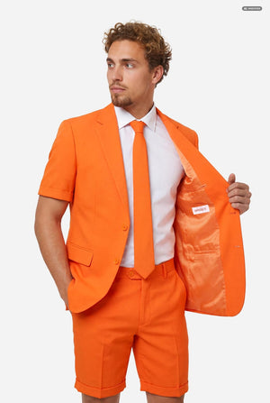 A man modeling a bright orange short-sleeve suit consisting of a blazer and matching shorts, paired with a white dress shirt and orange tie, against a light gray background. Holding one side of the jacket open.