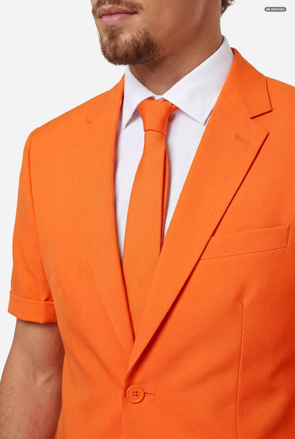 A man modeling a bright orange short-sleeve suit consisting of a blazer and matching shorts, paired with a white dress shirt and orange tie, against a light gray background. Chest close-up.