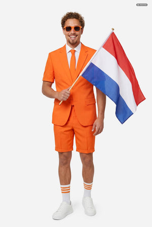A man modeling a bright orange short-sleeve suit consisting of a blazer and matching shorts, paired with a white dress shirt and orange tie, against a light gray background. Waving red, white and blue Dutch flag.