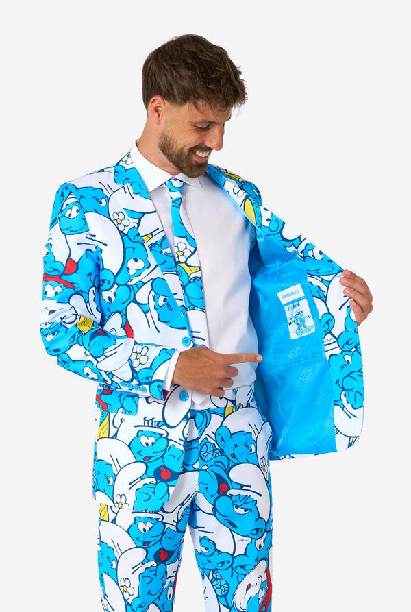 Smiling man wearing a colorful Smurfs men's suit with an all-over cartoon print in blue, white, red, and yellow, including a matching tie and white shirt, standing against a plain background. Holding one side of the jacket open.