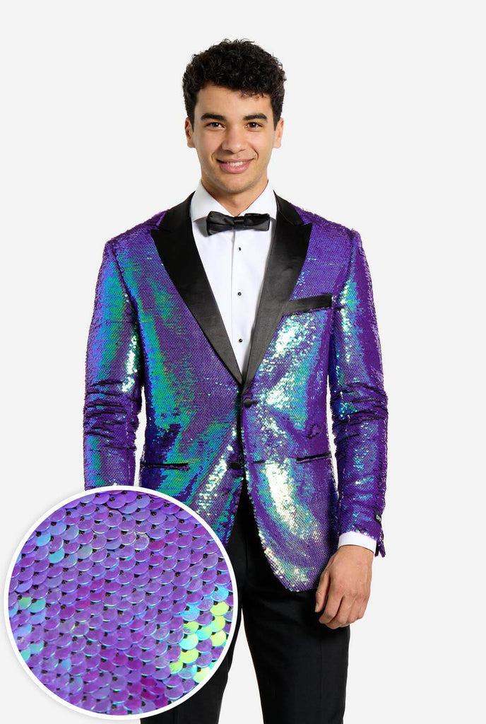 In this image, a man is dressed in a formal tuxedo featuring a shimmering, sequined blazer. The blazer has an iridescent finish, shifting between shades of blue, green, and purple, giving a high-gloss, reflective look. The jacket has black satin lapels and pockets, which stand out against the bright colors. He is wearing a crisp white dress shirt with black buttons and a black bow tie, paired with black formal trousers. The man is smiling and standing with a relaxed posture. Including close up circle.