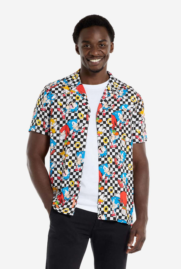 A man is wearing a black and white checkered Hawaiian shirt with characters from the Sonic the Hedgehog franchise and golden rings on it. Also wearing black pants. He is smiling in the camera.