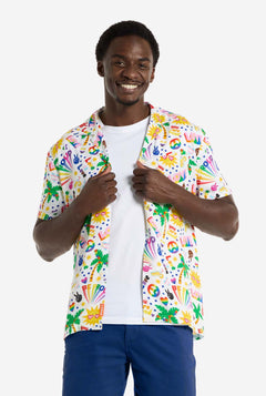 A man is wearing a white Hawaiian shirt with a rainbow pride print, Print consists of peace symbols, hearts and other icons, Also, wearing blue pants. He is smiling in the camera and holding the sides of the shirts.
