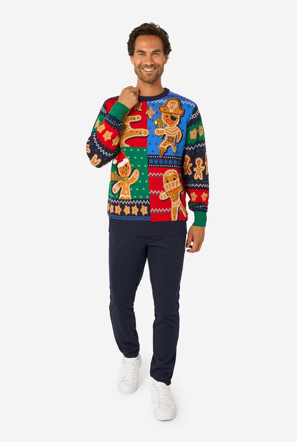 Full-body view of a man in a knitted Christmas sweater with gingerbread characters, navy pants, and white sneakers, smiling and posing.