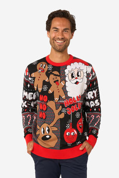 A man modeling a black Christmas crewneck sweater with colorful holiday graphics including gingerbread men, Santa, a reindeer, and 'Holly, No, Holly' text, paired with navy pants against a light gray studio background.