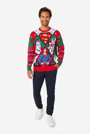 Full-body view of a man in a Superman-themed knitted Christmas sweater, navy trousers, and white sneakers.