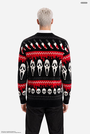 A male model with platinum blonde hair wearing a black and red Halloween-themed knit sweater with Ghostface and skull patterns, paired with black pants, against a light gray background. View from the back.