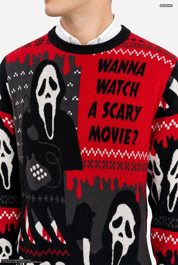 A male model with platinum blonde hair wearing a black and red Halloween-themed knit sweater with Ghostface and skull patterns, paired with black pants, against a light gray background. Chest close up.