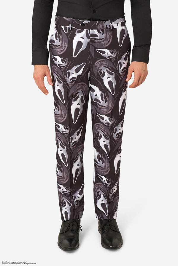 A person wearing black pants with an allover Ghostface mask print in white and gray, paired with a black long-sleeve top and black shoes, shown against a light gray studio background.