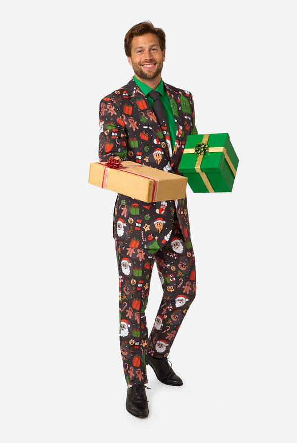 A man poses in a black background colored christmas suit decorated with festive holiday imagery like reindeer, snowflakes, and gift boxes. Holding two presents