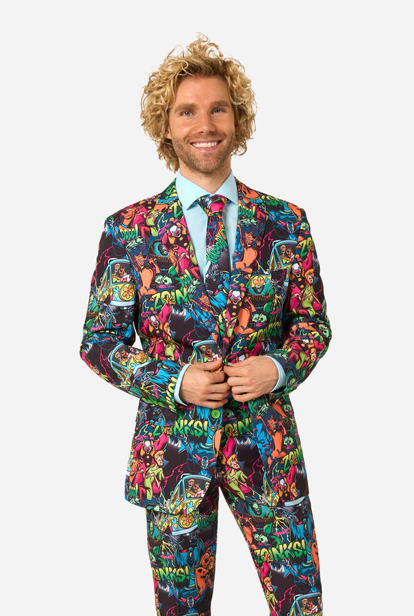 A man with curly blond hair wearing a vibrant, multicolored Scooby-Doo-patterned suit, a light blue dress shirt, and a matching tie, standing against a solid light gray background.