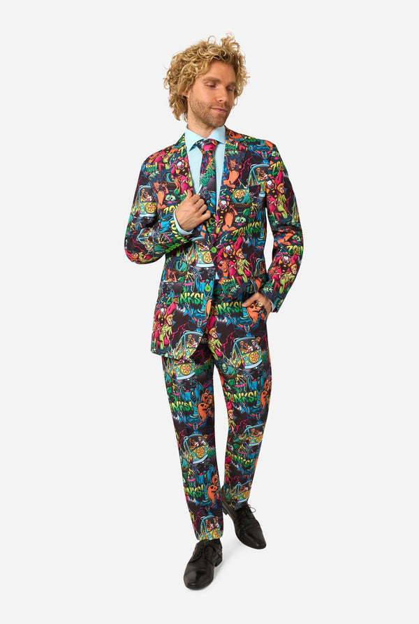 A man modeling a vibrant, multicolored Scooby-Doo suit with a busy graffiti-style print, paired with a light blue dress shirt and a patterned tie, against a plain white background.
