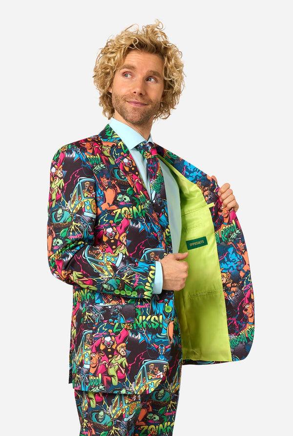 A man with curly blond hair models a vibrant, multicolored Scooby-Doo style suit jacket and matching trousers, holding open the jacket to reveal a bright lime green interior lining. He is wearing a light blue dress shirt and a matching tie against a solid light gray background.