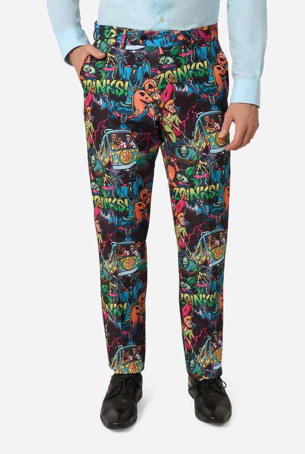 A person wearing black pants with a highly colorful, vibrant Scooby-Doo print featuring retro arcade machines, cartoon characters, and neon text, styled with a light blue dress shirt and black dress shoes against a plain light gray background.