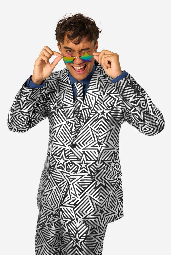 Man playfully smiling and adjusting reflective rainbow-colored star-shaped sunglasses while wearing bold geometric suit.