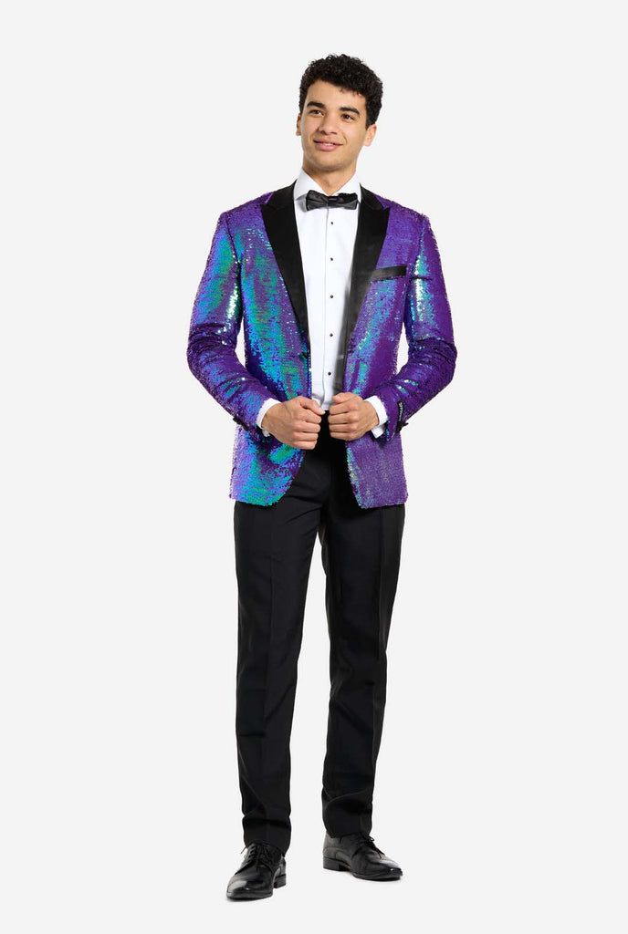 
In this image, a man is dressed in a formal tuxedo featuring a shimmering, sequined blazer. The blazer has an iridescent finish, shifting between shades of blue, green, and purple, giving a high-gloss, reflective look. The jacket has black satin lapels and pockets, which stand out against the bright colors. He is wearing a crisp white dress shirt with black buttons and a black bow tie, paired with black formal trousers. The man is smiling and standing with a relaxed posture, hands holding the jacket.