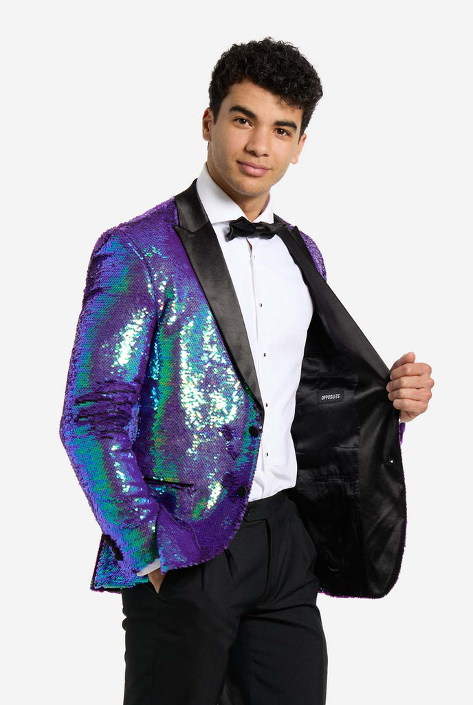 
In this image, a man is dressed in a formal tuxedo featuring a shimmering, sequined blazer. The blazer has an iridescent finish, shifting between shades of blue, green, and purple, giving a high-gloss, reflective look. The jacket has black satin lapels and pockets, which stand out against the bright colors. He is wearing a crisp white dress shirt with black buttons and a black bow tie, paired with black formal trousers. The man is smiling and standing sideways one hand holding side of jacket.