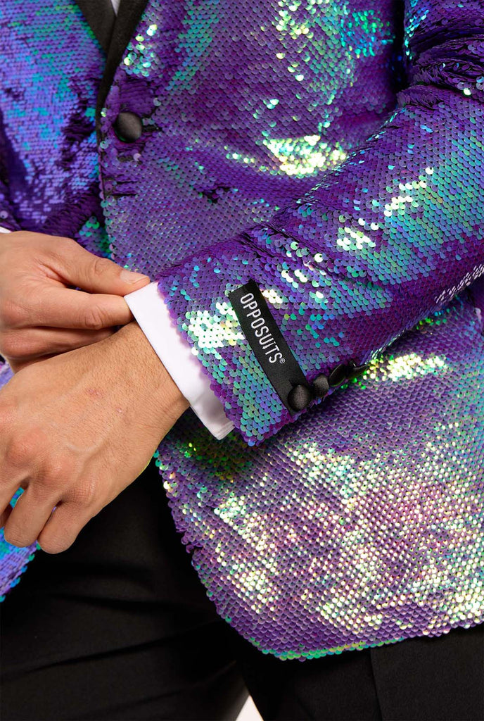 
In this image, a man is dressed in a formal tuxedo featuring a shimmering, sequined blazer. The blazer has an iridescent finish, shifting between shades of blue, green, and purple, giving a high-gloss, reflective look. The jacket has black satin lapels and pockets, which stand out against the bright colors. Close up.