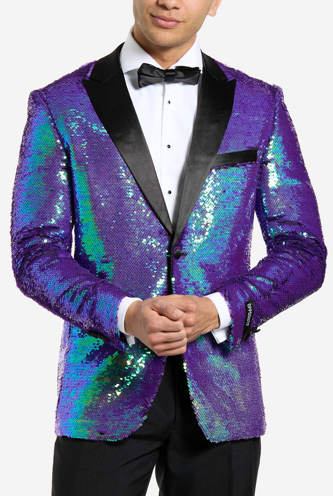 
In this image, a man is dressed in a formal tuxedo featuring a shimmering, sequined blazer. The blazer has an iridescent finish, shifting between shades of blue, green, and purple, giving a high-gloss, reflective look. The jacket has black satin lapels and pockets, which stand out against the bright colors. He is wearing a crisp white dress shirt with black buttons and a black bow tie, paired with black formal trousers. Chest close up.