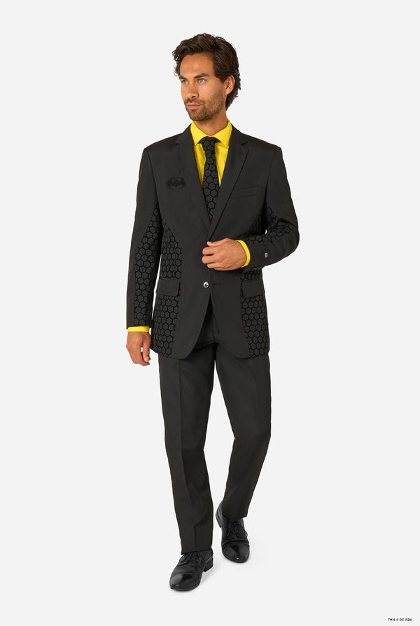 Full-body view of a man wearing a black Batman suit with a yellow shirt and black tie, standing at a slight angle on a white background.