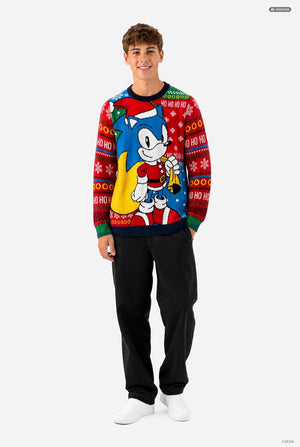 A young man modeling a festive holiday crewneck sweater featuring Sonic the Hedgehog on a red Christmas patterned background, paired with black sweatpants and white sneakers against a light gray studio backdrop.