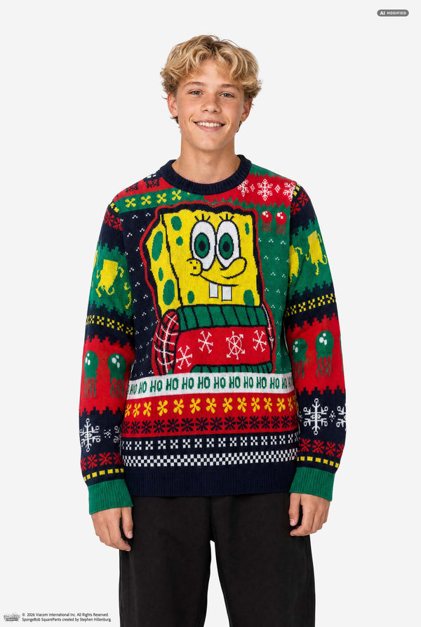 A young man wearing a SpongeBob-themed Christmas sweater, standing against a plain light gray studio background. The sweater features a large SpongeBob graphic on the chest and festive multicolor holiday patterns including snowflakes, bells, and 'HO HO HO' text.